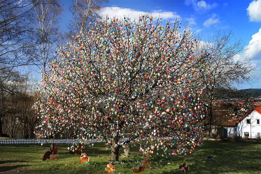A large tree decorated with numerous colorful Easter eggs stands in an open area, with small decorative bunnies placed around its base.