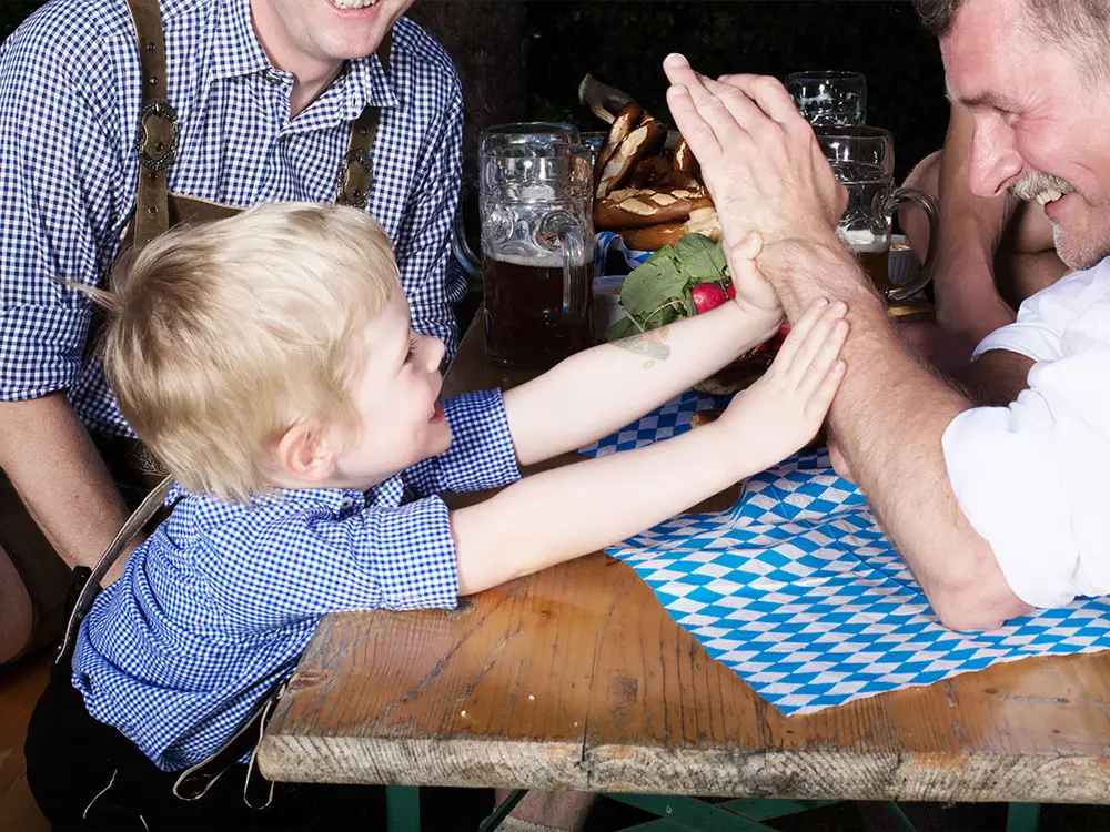 Young boy in traditional German clothing playfully arm wrestling an adult at an Oktoberfest beer table.
