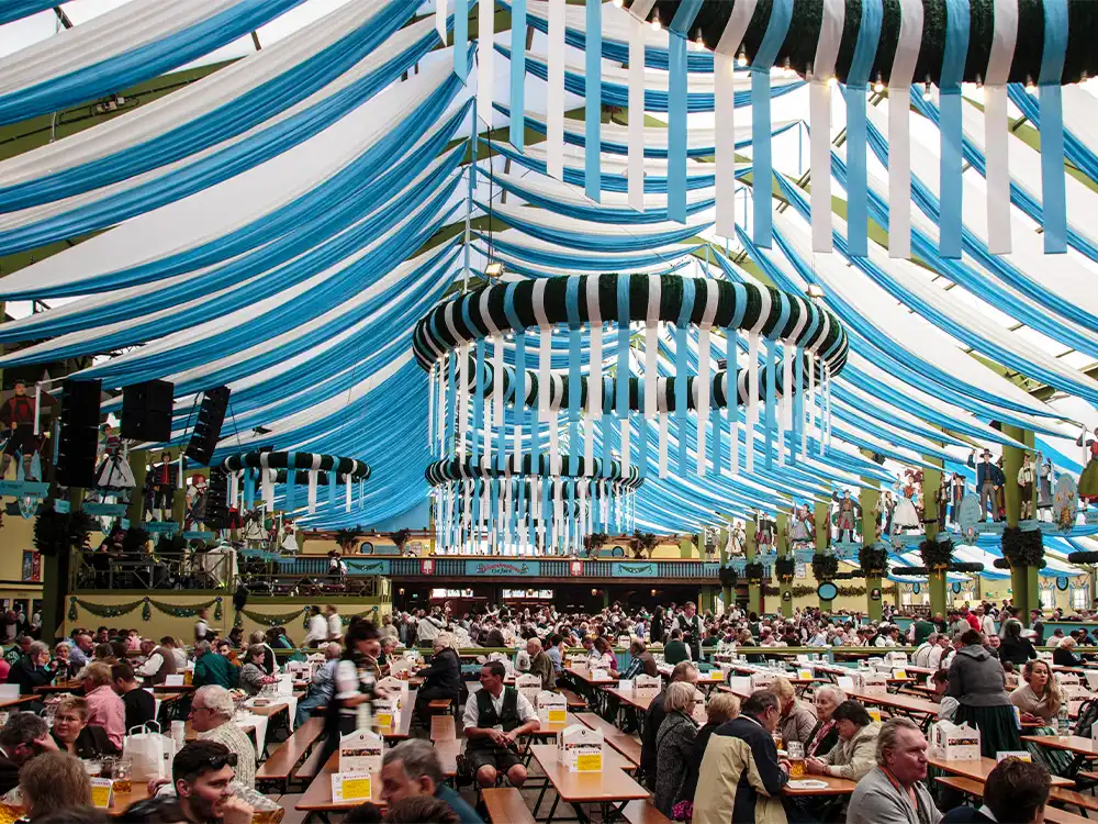 Interior of a traditional Oktoberfest beer tent decorated with blue and white ribbons and full of seated festivalgoers.