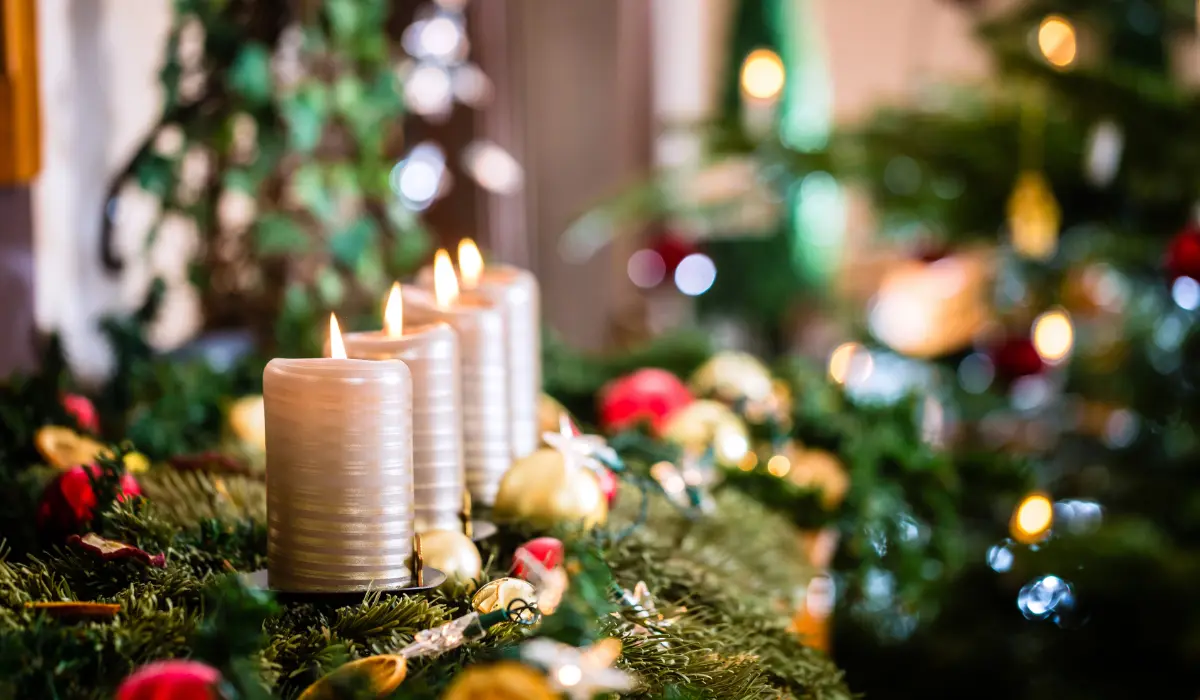 Four lit candles on a festive wreath with greenery, red and yellow ornaments, and blurred lights in the background.