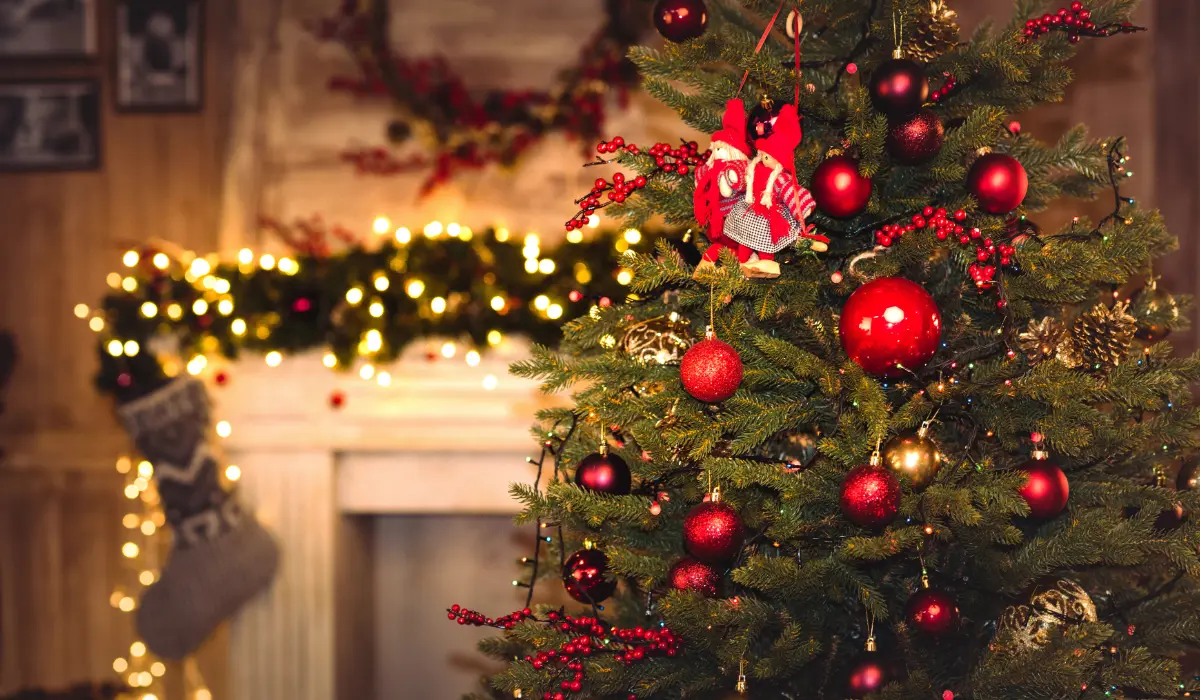 A decorated Christmas tree with red and gold ornaments stands in a warmly lit room. A fireplace adorned with garlands and stockings is visible in the background.