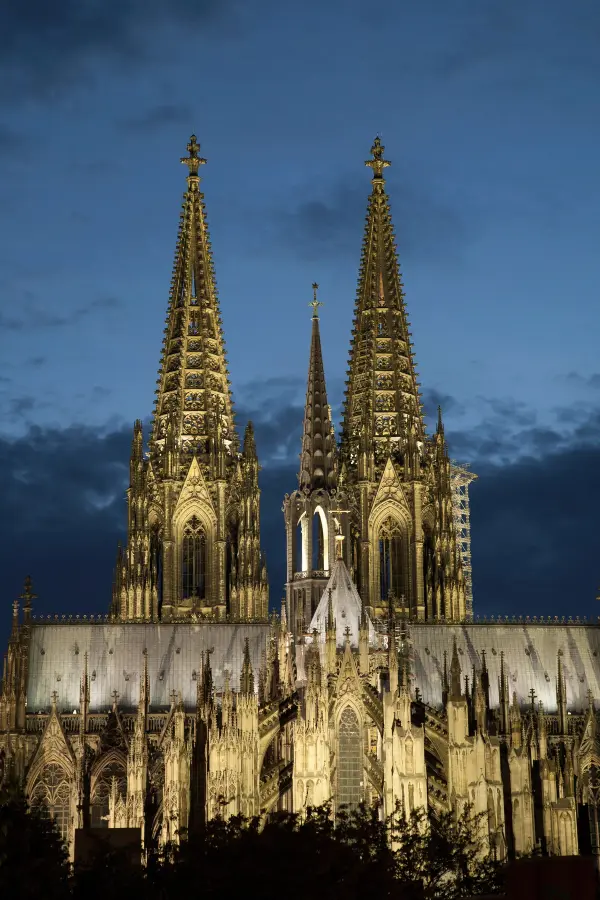 Night view of the Gothic-style Cologne Cathedral with twin spires and intricate architectural details, illuminated against a dark blue evening sky.