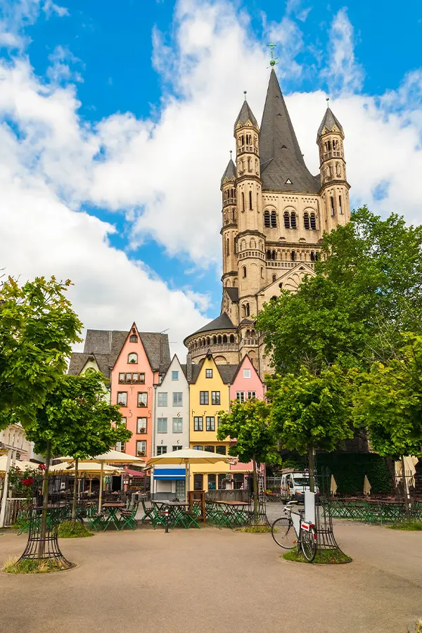 A historic church tower with rounded spires stands behind colorful, gabled buildings in a tree-lined square with outdoor seating and parked bicycles under a cloudy blue sky.