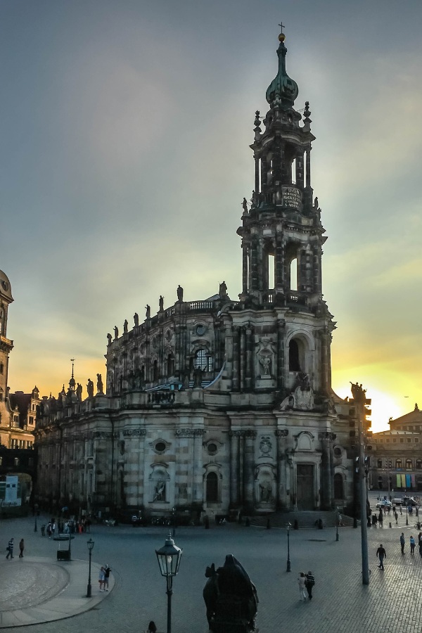 The ornate, historic Dresden Cathedral with statues, arched windows, and a tall clock tower stands in a city square at sunset, with people scattered around.