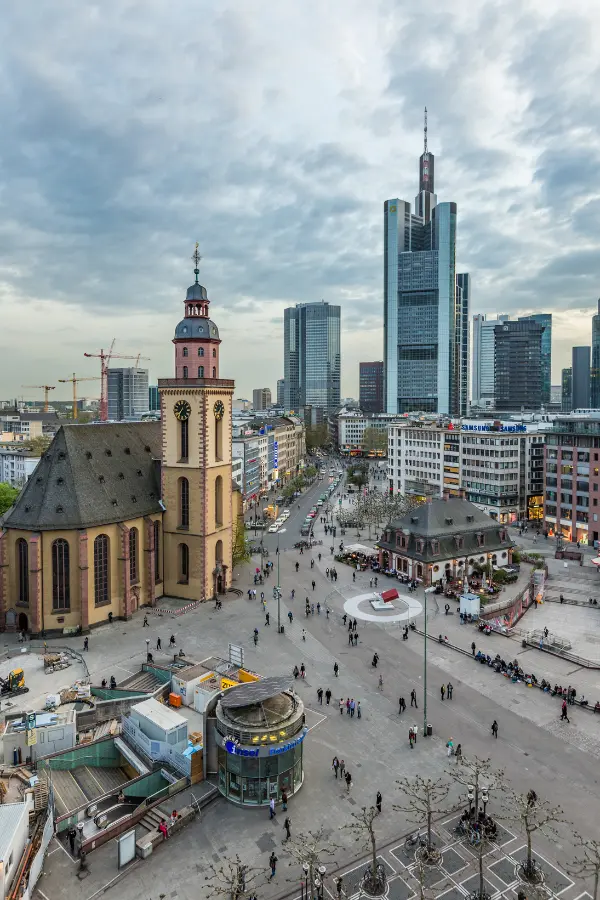 An elevated view of Frankfurt, showcasing a mix of historic and modern architecture with St. Catherine's Church to the left and high-rise buildings in the background. People fill the plaza below.