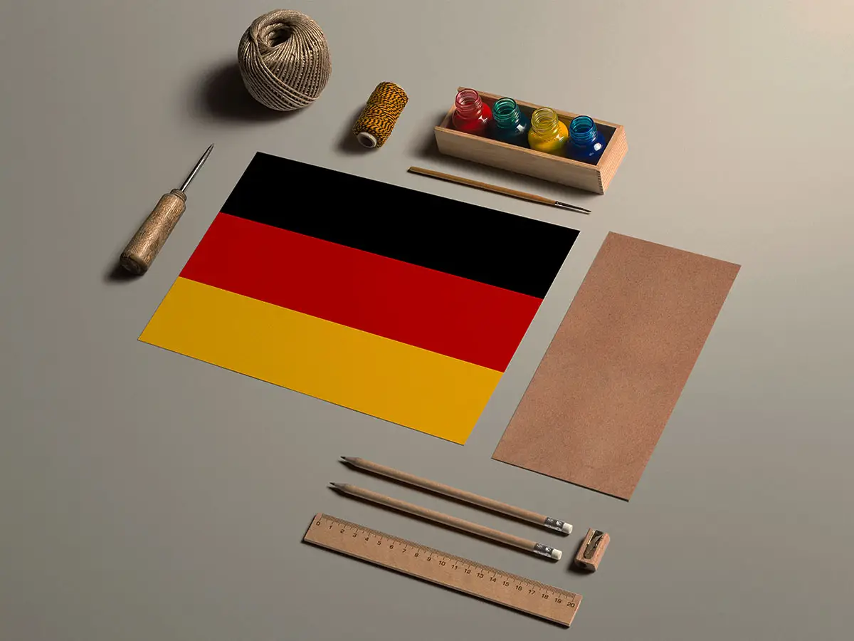 Flatlay of craft tools surrounding a German flag, including spools of thread, a ruler, paint jars, and wooden pencils arranged neatly on a beige surface.