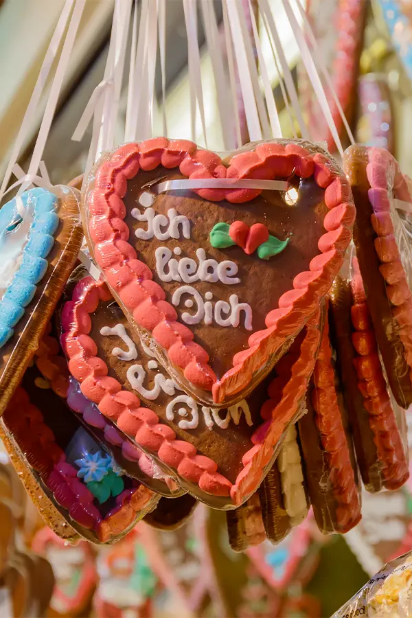 Heart-shaped gingerbread cookies with pink icing borders hang on display. One cookie features German text "Ich liebe Dich" and a red rose decoration.