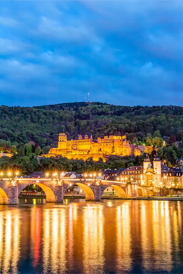 A lit-up Heidelberg Castle on a hill with a stone bridge reflecting in the river below, set against a backdrop of trees and a cloudy evening sky.