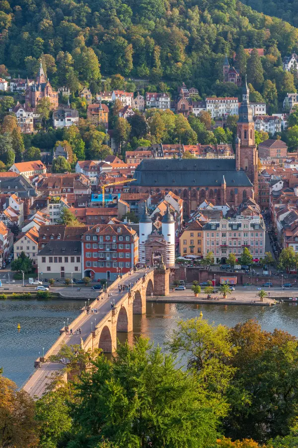 A panoramic view of Heidelberg, Germany, featuring the Old Bridge, the Church of the Holy Spirit, and surrounding buildings, set against a backdrop of lush, green hills.