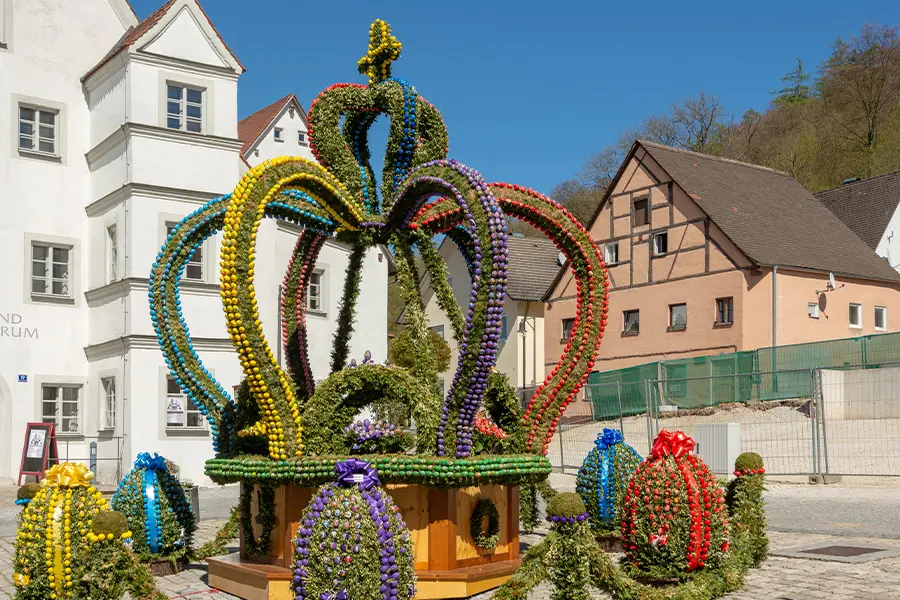Large, colorful crown sculpture made of painted eggs stands in a sunny courtyard, surrounded by traditional buildings and greenery in the background.