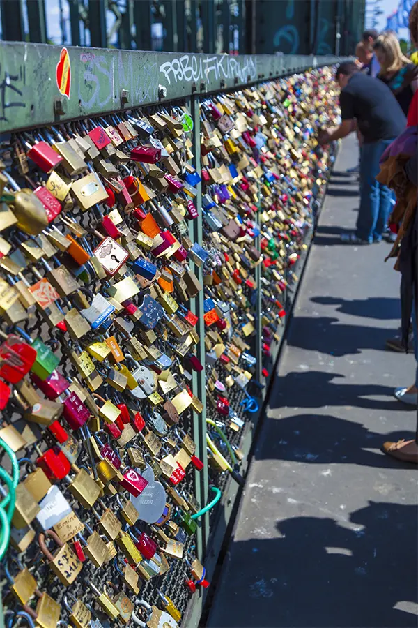 A railing covered with numerous colorful padlocks, with several people standing nearby on a sunny day.