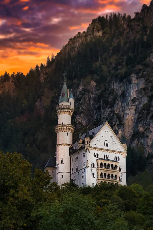 Neuschwanstein castle with multiple towers and pointed roofs stands in front of a rugged mountainside with trees, under a brightly colored sunset sky.