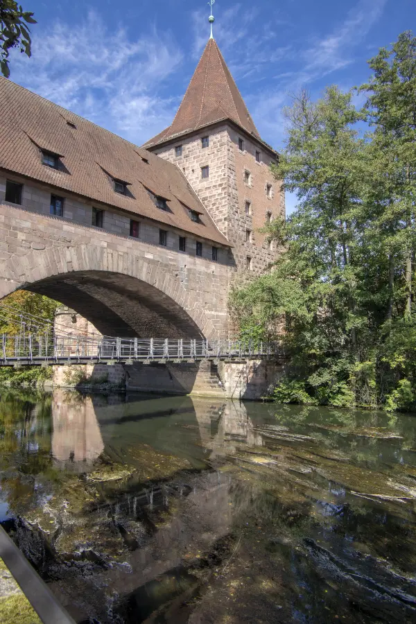 A medieval stone tower and arch bridge over a calm river, with trees and buildings nearby, under a clear blue sky in Nuremberg, Germany.