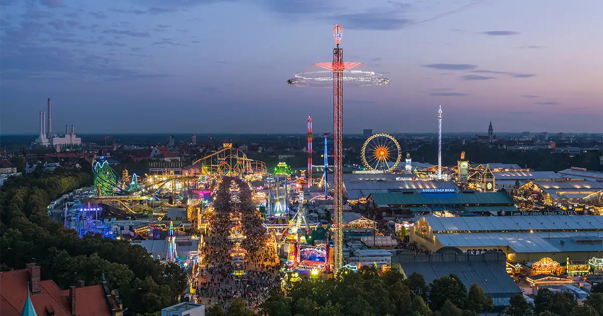 Nighttime aerial view of the Oktoberfest fairgrounds lit up with colorful lights, ferris wheels, rides, and bustling crowds.
