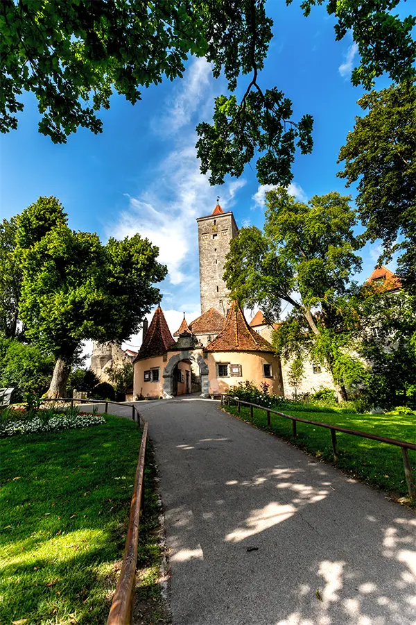 A paved path leads to a stone tower with a red roof in Rothenburg, surrounded by lush green trees under a blue sky.