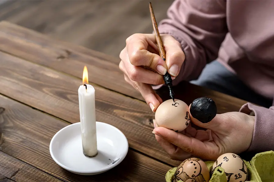 Person decorating eggs with wax near a lit candle on a wooden table.