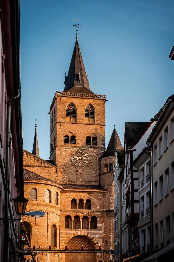 A tall, historic stone cathedral with multiple spires and a clock on its facade, flanked by buildings in a narrow street, illuminated by warm sunlight in Trier, Germany.