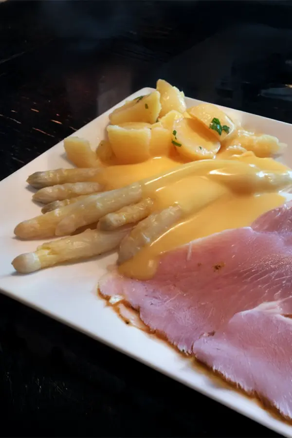 Plate with sliced ham, white asparagus covered in hollandaise sauce, and boiled potatoes garnished with herbs.