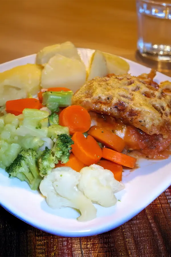A plate with cooked vegetables, including broccoli, carrots, and cauliflower, alongside baked potatoes and a piece of baked fish. A glass of water is in the background.