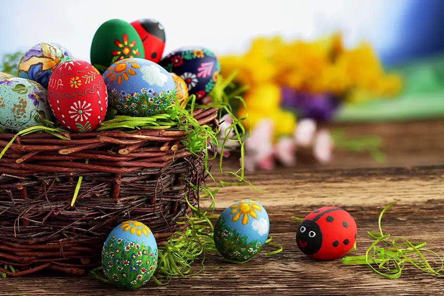 A basket of decorated Easter eggs on a wooden surface, with additional painted eggs, including a ladybug design, scattered outside. Yellow flowers are blurred in the background.