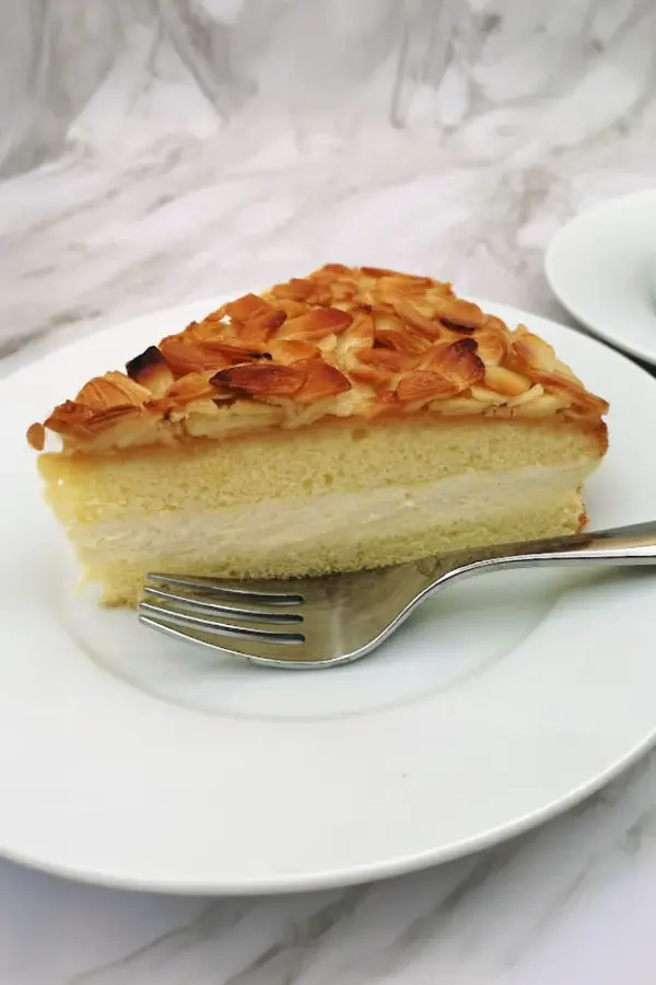 A slice of almond-topped bee sting cake with cream filling on a white plate, and a fork beside it.
