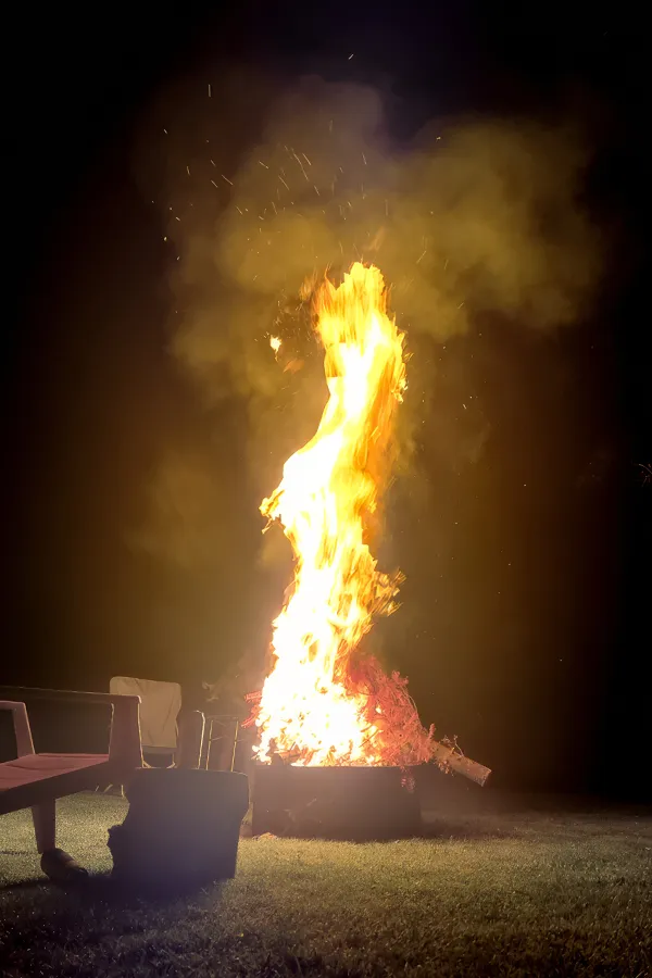 A large bonfire blazes brightly at night, surrounded by chairs on a grassy area.
