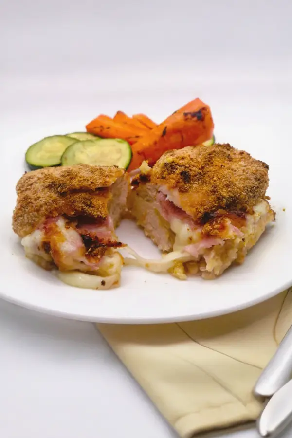 Sliced breaded chicken stuffed with ham and cheese on a white plate, accompanied by grilled zucchini and carrots.