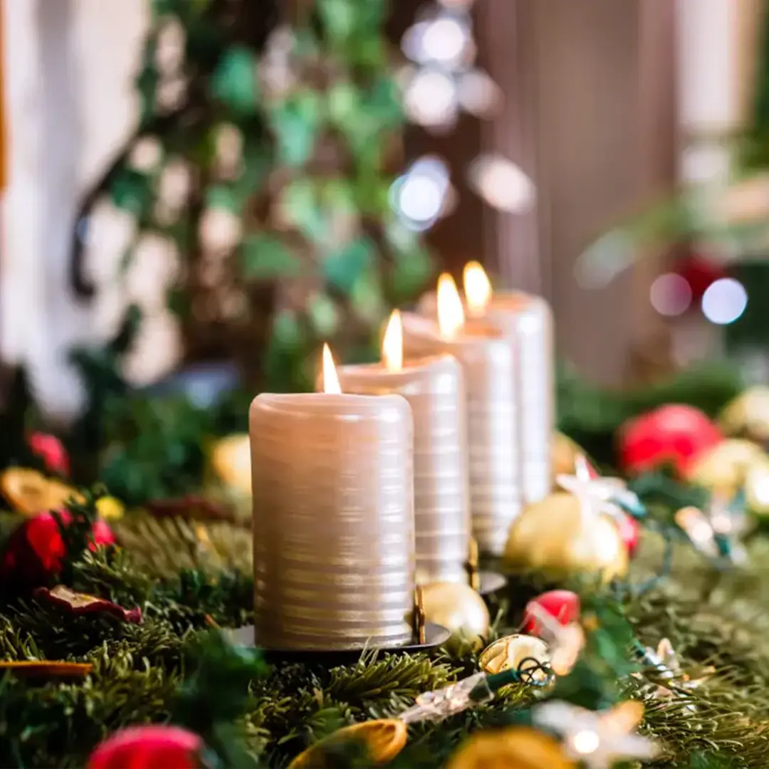 Four lit candles on a festive Advent wreath decorated with evergreen branches, red and gold ornaments, and twinkling lights in a cozy holiday setting.