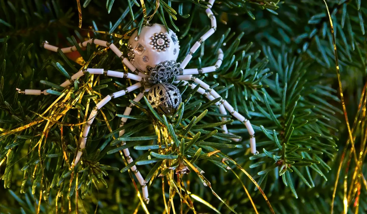 A decorative spider ornament made of beads and metal gears is placed on a Christmas tree, surrounded by green pine needles and golden tinsel.