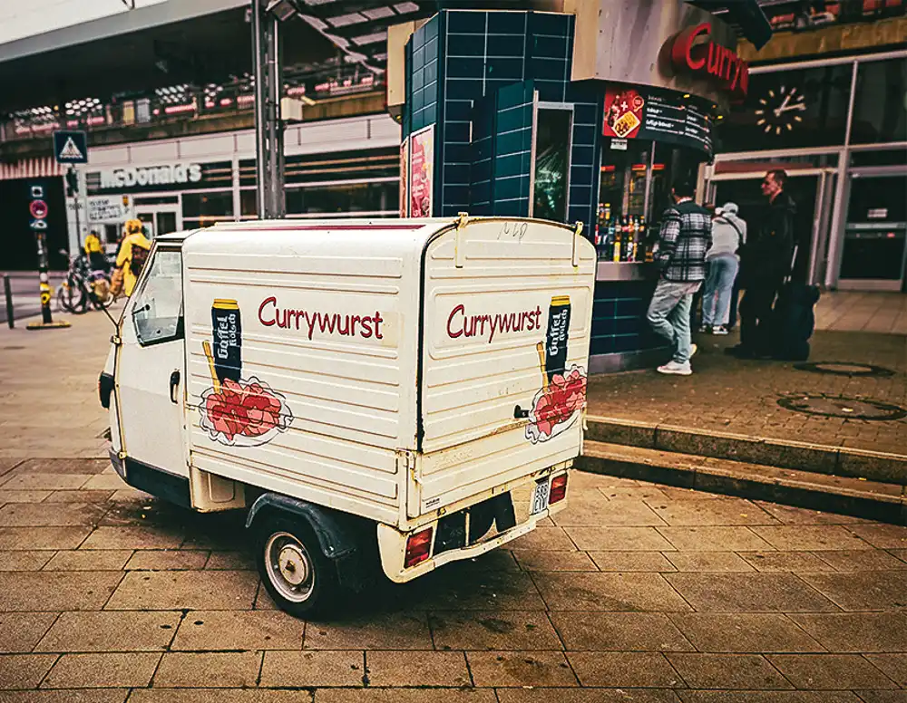 Small white delivery van advertising currywurst parked near a German street food stand.