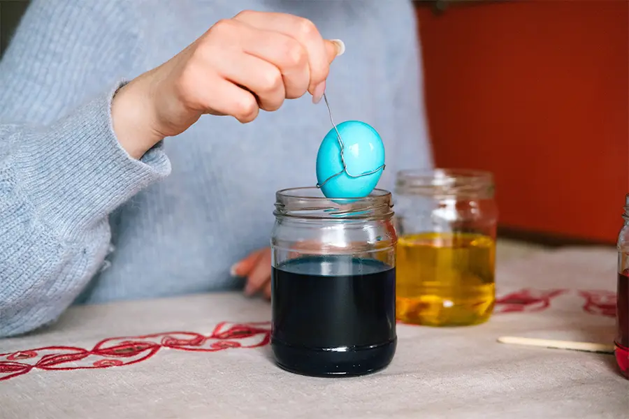 A person dyes an egg blue by dipping it in a jar of blue liquid.