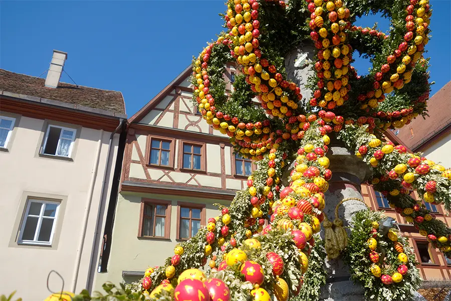 Fountain decorated with colorful Easter eggs and greenery in front of traditional timber-framed houses under a clear blue sky.