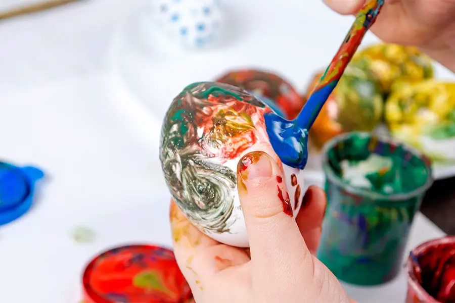 Child painting an egg with various colors using a paintbrush, surrounded by pots of paint.