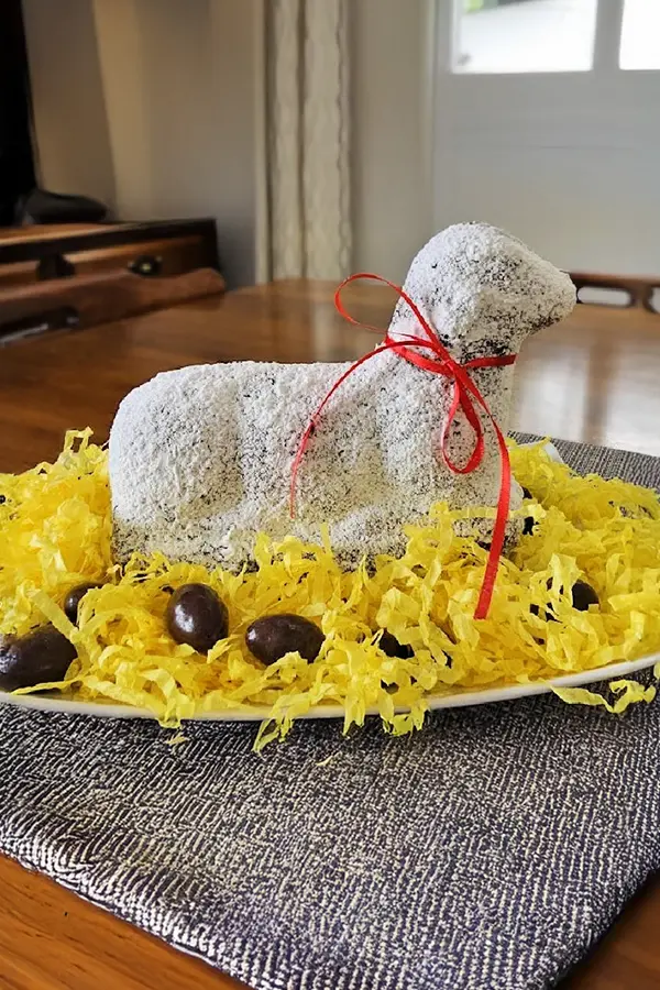Lamb-shaped cake covered in powdered sugar with a red ribbon, surrounded by yellow shredded paper and chocolate eggs on a plate.