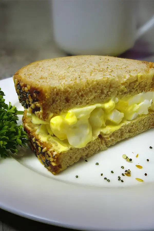 Egg salad sandwich on seeded bread, garnished with parsley on a white plate.
