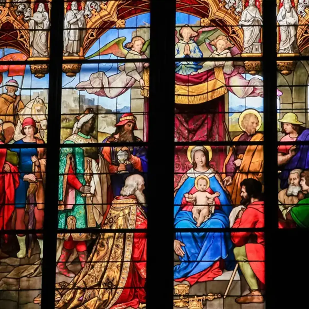 A vibrant stained glass window depicting the biblical scene of the Three Wise Men presenting gifts to baby Jesus, surrounded by colorful figures and ornate details.