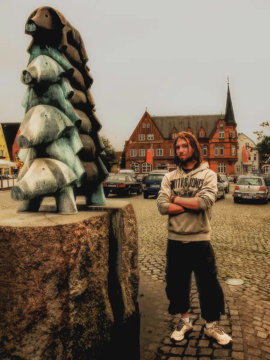 Man stands confidently beside a whimsical stacked pig statue in a Hamburg town square, with cobblestone streets and red-brick buildings in the background.