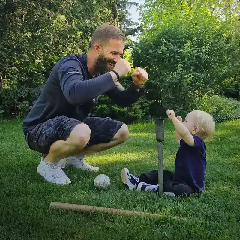 Bearded man squats and playfully cheers on a toddler holding a baseball bat, sitting on a lawn with scattered baseball gear.
