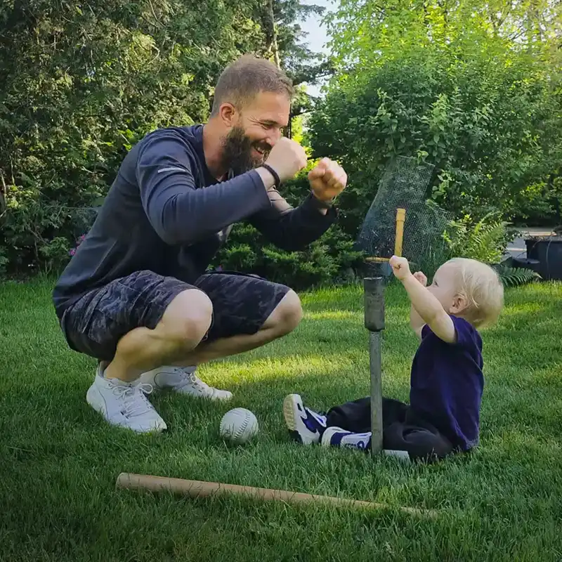 Bearded man squats and playfully cheers on a toddler holding a baseball bat, sitting on a lawn with scattered baseball gear.