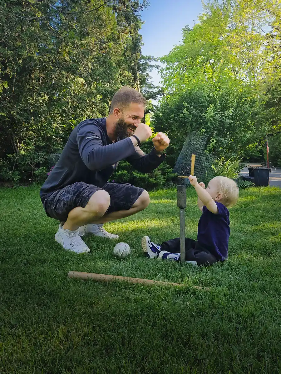 Bearded man squats and playfully cheers on a toddler holding a baseball bat, sitting on a lawn with scattered baseball gear.