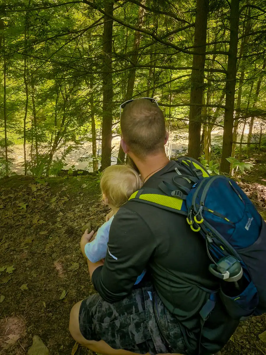 A man wearing a black shirt and backpack kneels beside a toddler in a wooded forest, both facing away and looking into the trees.