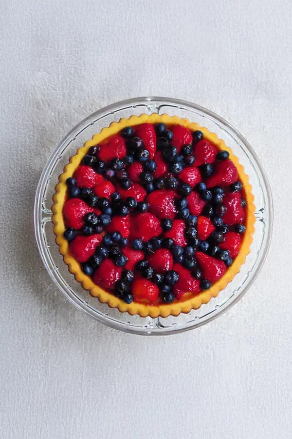 A round fruit tart with a yellow crust, topped with strawberries and blueberries, on a glass plate against a light background.