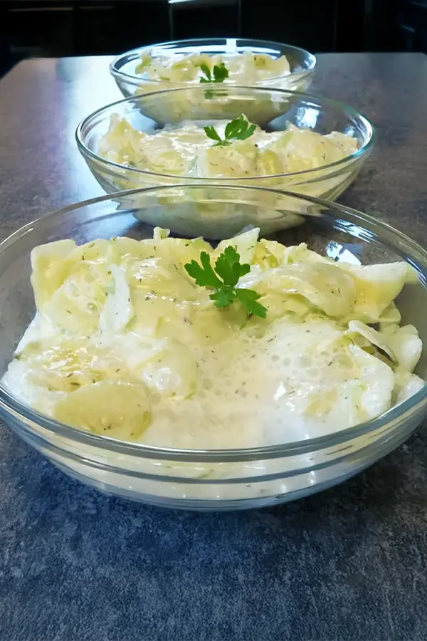 Three glass bowls of creamy cucumber salad topped with parsley are arranged in a line on a gray countertop.