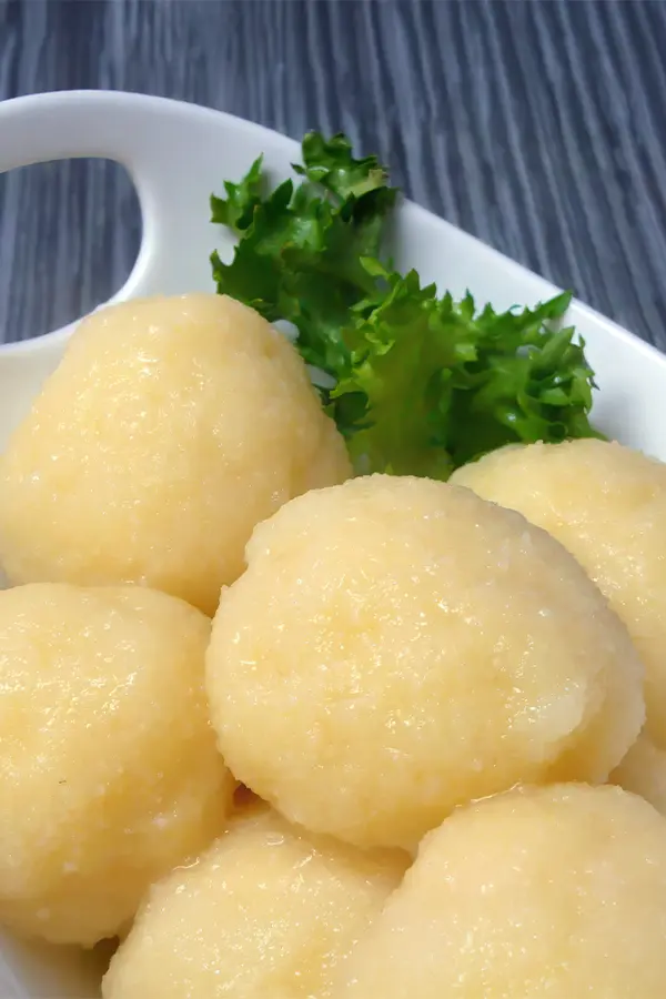 Close-up of six round dumplings in a white dish with a piece of green leafy garnish, placed on a gray wooden surface.