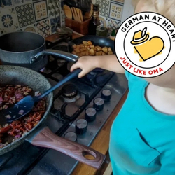 A blond toddler stirs a pan of onions and sun-dried tomatoes at the stove, learning German cooking by helping rather than being taught.