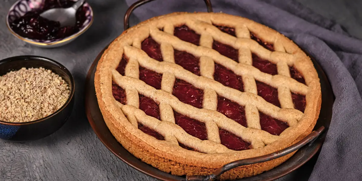 Traditional Linzertorte with a lattice crust and red jam filling, served with a bowl of ground nuts and jam.