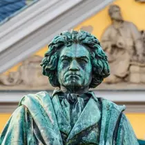 A bronze statue of composer Ludwig van Beethoven with a stern expression, set in front of a yellow historic building in Bonn, Germany.