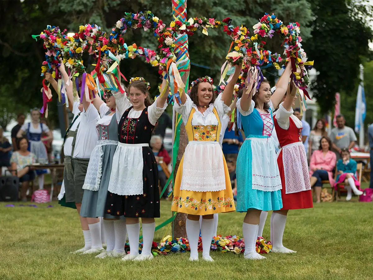 A group of young women in traditional German dresses (dirndls) raise decorated wreaths above their heads during a lively maypole dance at an outdoor German festival.