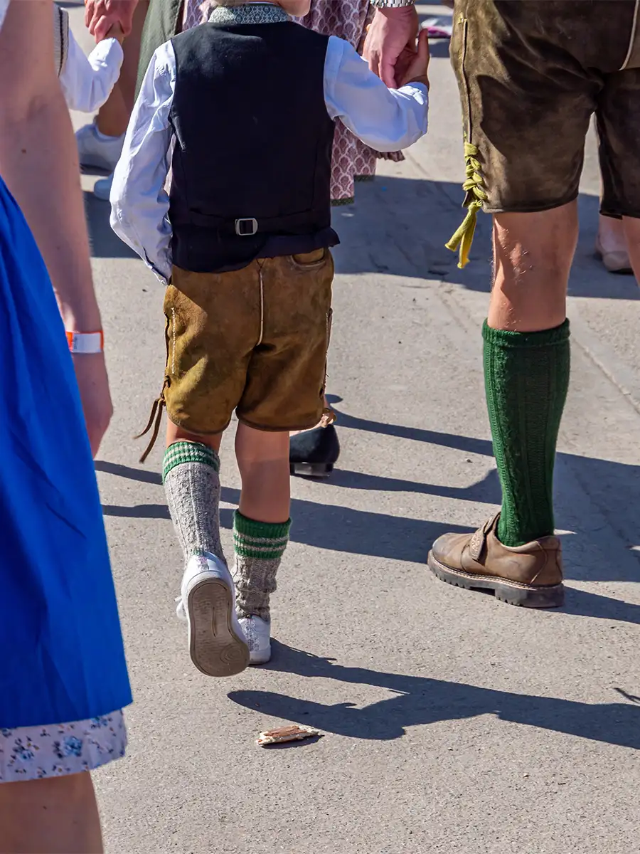 Back view of a young boy and man wearing lederhosen and traditional socks, walking among a crowd at Oktoberfest.