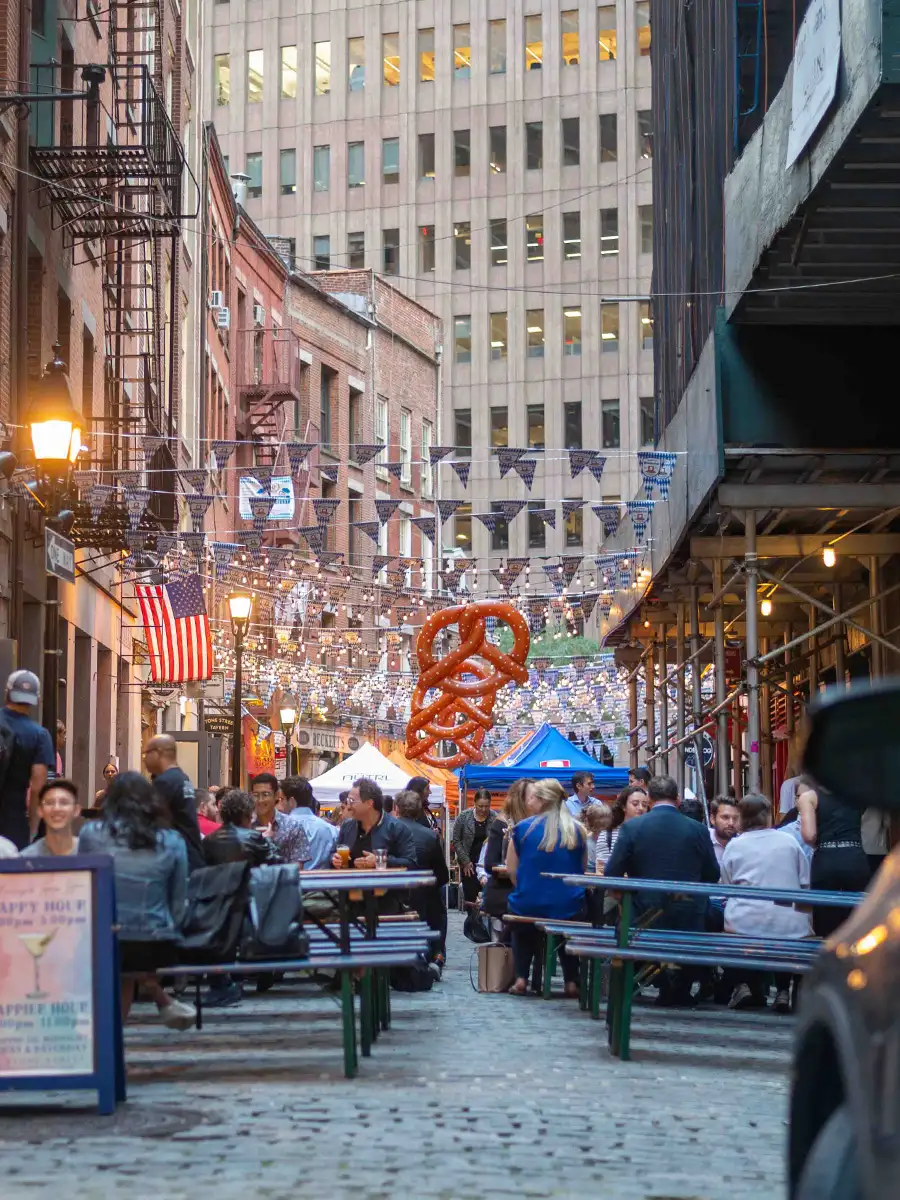 Street view of an Oktoberfest celebration in New York with long tables, crowds, American flags, and a giant pretzel decoration overhead.